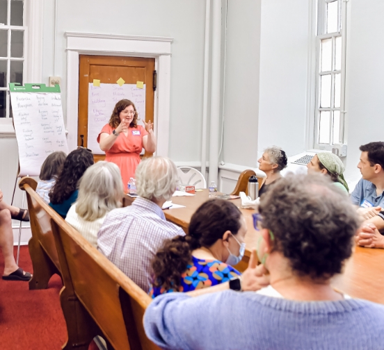 An intergenerational group of people sitting at a long table and listening to a woman who stands at the head of the table. She is wearing a beach dress and has her hands held in front of her as if gesturing while speaking. She has two large papers around her on an easel and taped to the wall behind her. 