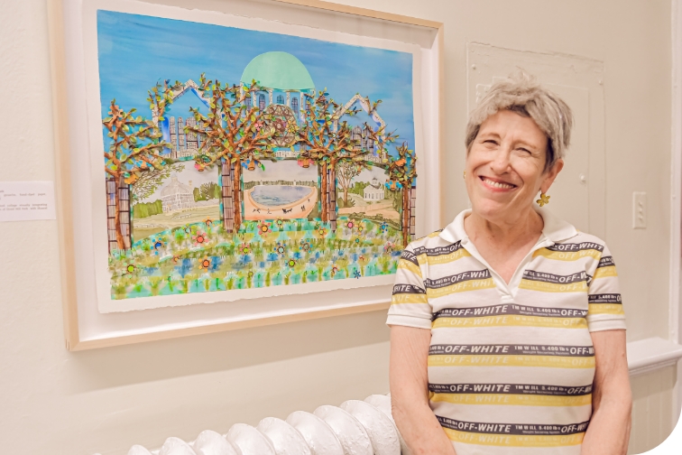 A person with short hair, visible from the mid-torso up and wearing a short sleeved striped shirt with a collar smiles at the camera in front of a framed collage depicting a green dome behind trees.