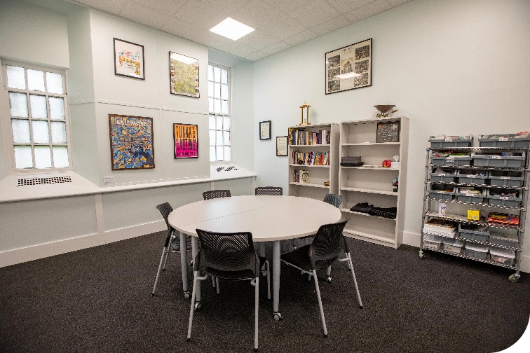 Conference room with movable round table and five chairs, book shelves, wire shelves with office supplies, and framed posters on the wall.