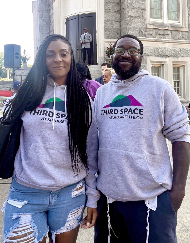 A woman and a man, both visible from mid-thigh up and wearing sweatshirts that read Third Space at Shaarei Tfilloh, stand in front of the Third Space building. The woman has long braids that fall on either shoulder. She is wearing denim shorts with unraveling hems. The man has dark hair, a beard, and wears glasses. He is wearing dark pants, and his tzitzit (ritual fringe) is visible below his sweatshirt. 