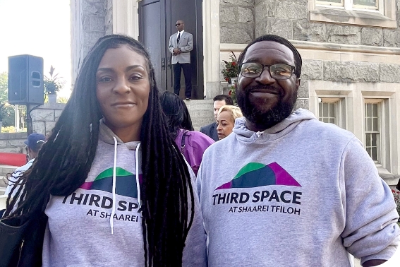 A woman and a man, both visible from mid-torso up and wearing sweatshirts that read Third Space at Shaarei Tfilloh, stand in front of the Third Space building. The woman has long braids that fall on either shoulder. The man has dark hair, a beard, and wears glasses.