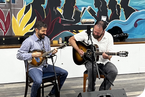 Two men sit in front of microphones playing instruments. The man on the left plays a banjo and smiles toward the right of the frame. The man on the right plays a guitar and looks down toward his instrument. Behind them is a stage and a mural in blues, blacks, yellow, and magenta.