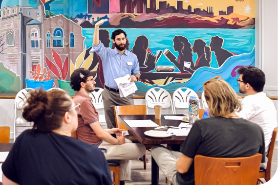 Four people sit around a table facing a man at the front of the room who is standing. The standing man has dark hair and a beard. He wears a blue dress shirt and holds one hand above his head and parallel to the ground, in the other hand he holds papers. 