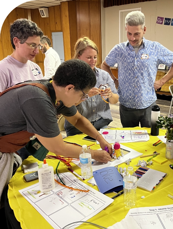 Four people standing around a table, working on a small sculpture. In the foreground, someone in a gray shirt and brown overalls with short dark hair and glasses leans over to adjust something on the sculpture. There are craft supplies strewn about on the table. Three other people look on. 