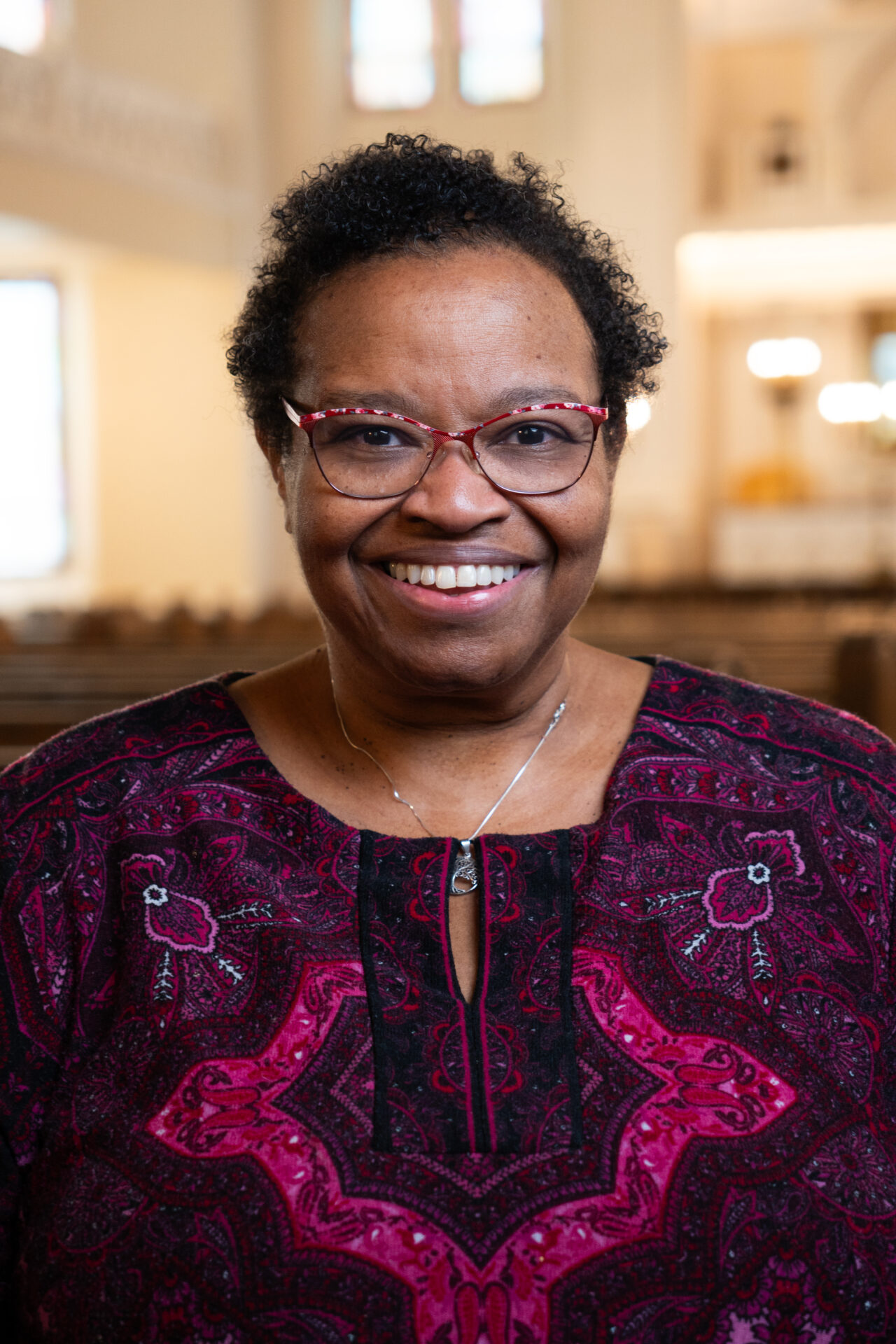 Kohenet Dr. Harriette Wimms, a brown skinned woman with short curly hair and glasses, from the mid torso up, smiling at the camera, wearing a purple and magenta embroidered tunic