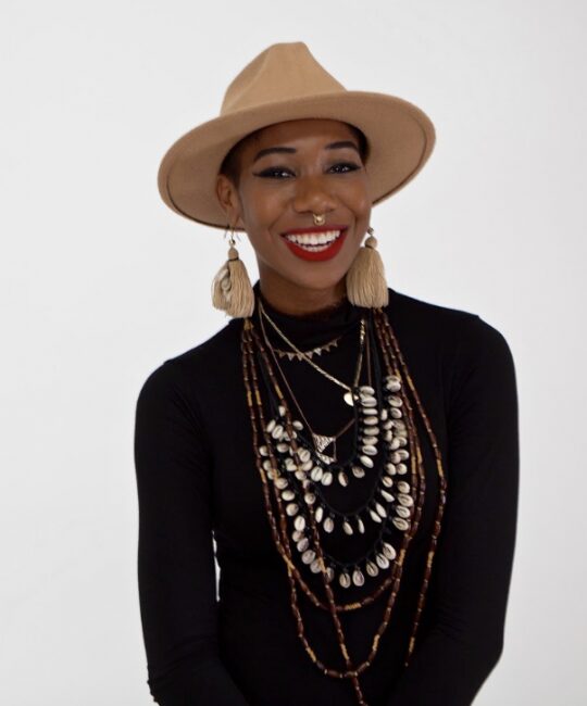 Dazia Wallerson, a brown skinned woman wearing a light brown hat and big earrings, from the mid-torso up, smiling at the camera, wearing a black turtleneck and multiple beaded necklaces