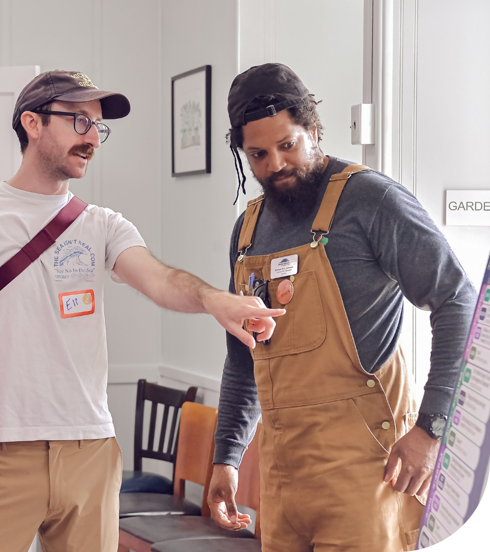 A man with glasses and mustache points to something outside the frame of the photo. Next to him, a bearded man in work overalls and with a few braids loose around his face looks in the direction of his companions pointing finger. 