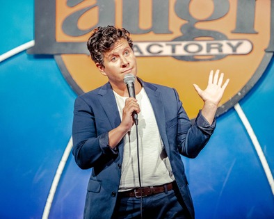 Liz Glazer wearing a tee shirt and blue blazer, holding a microphone to her mouth and gesturing with the other hand