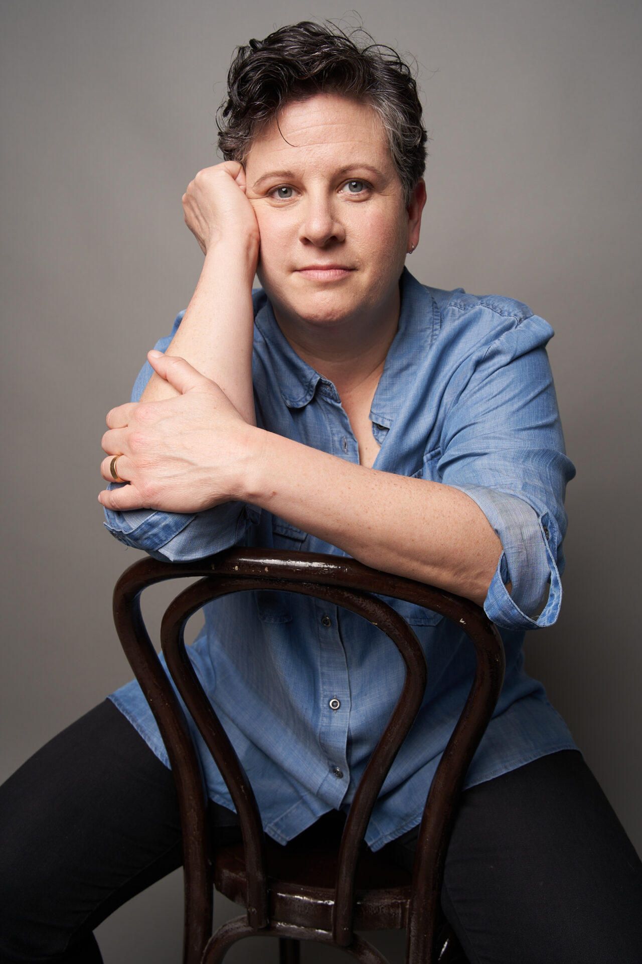 Liz Glazer, a fair skinned person with short hair, wearing a blue button-up shirt and dark pants, sits backward on a chair, facing the camera. One hand is fisted next to her cheek, the other rests on her forearm.