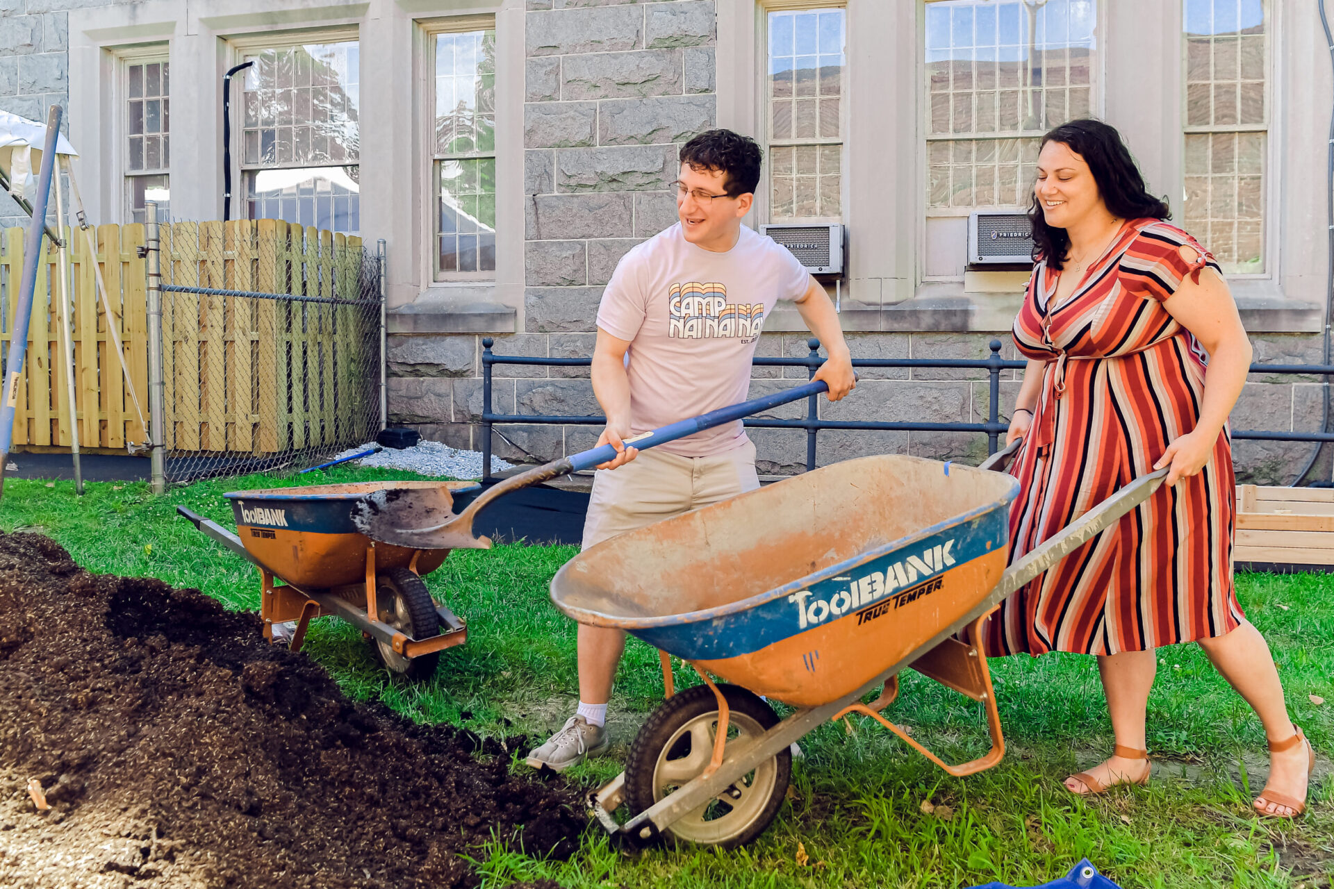 Two people, one in tee-shirt and shorts holding a shovel, the other wearing a striped dress and lifting the handles of a wheel barrow smile as they move soil.