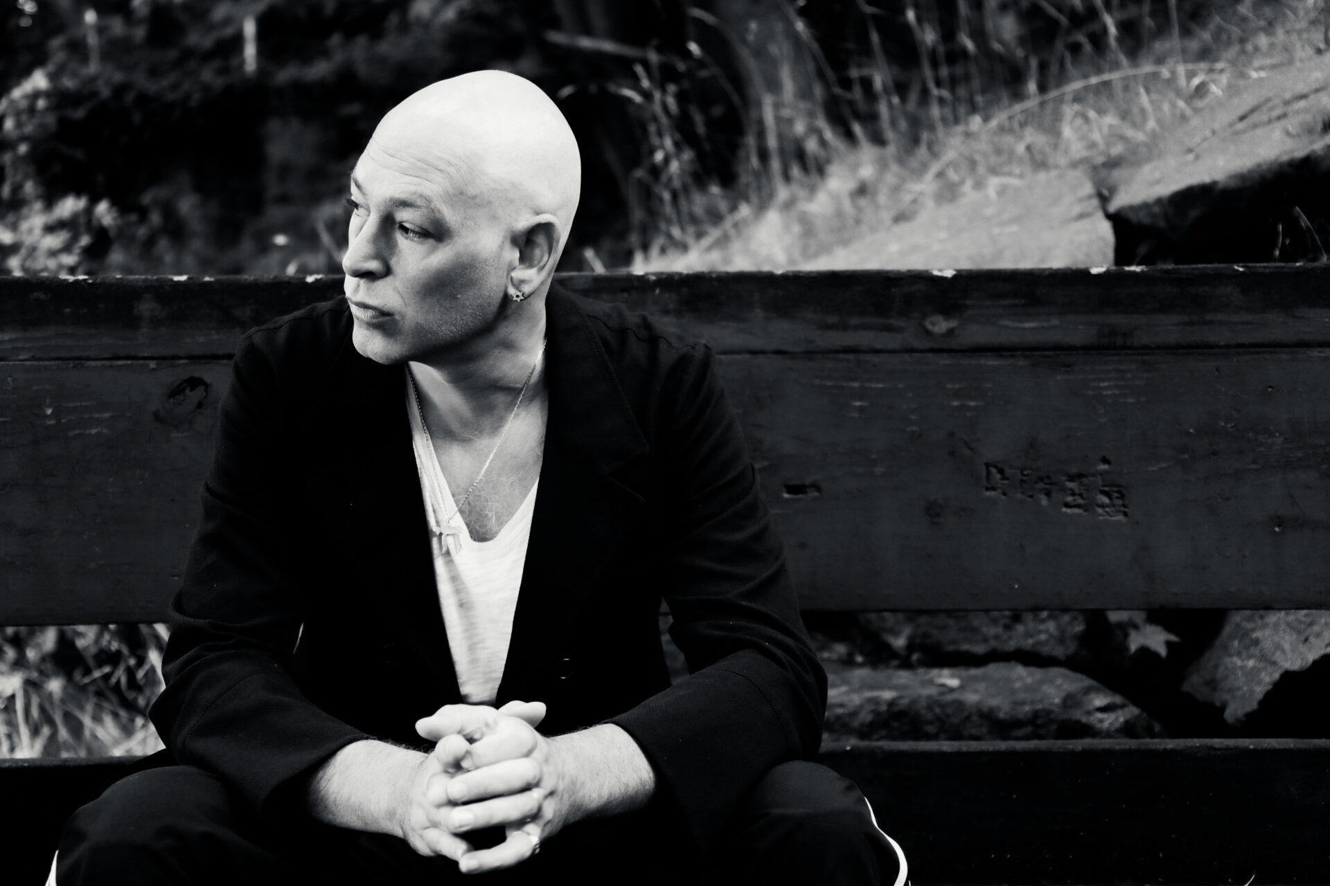 Black and white photo of Matisyahu with close cropped hair, wearing a white teeshirt and dark blazer, seated and looking off to the left.