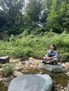 A person dressed in brown pants and a plaid shirt open over a tee shirt seated cross-legged beside a river