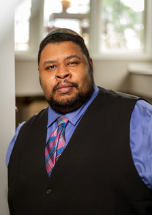 Michael Twitty wearing periwinkle dress shirt, plaid tie, and dark vest, from the mid-torso up, looking at the camera