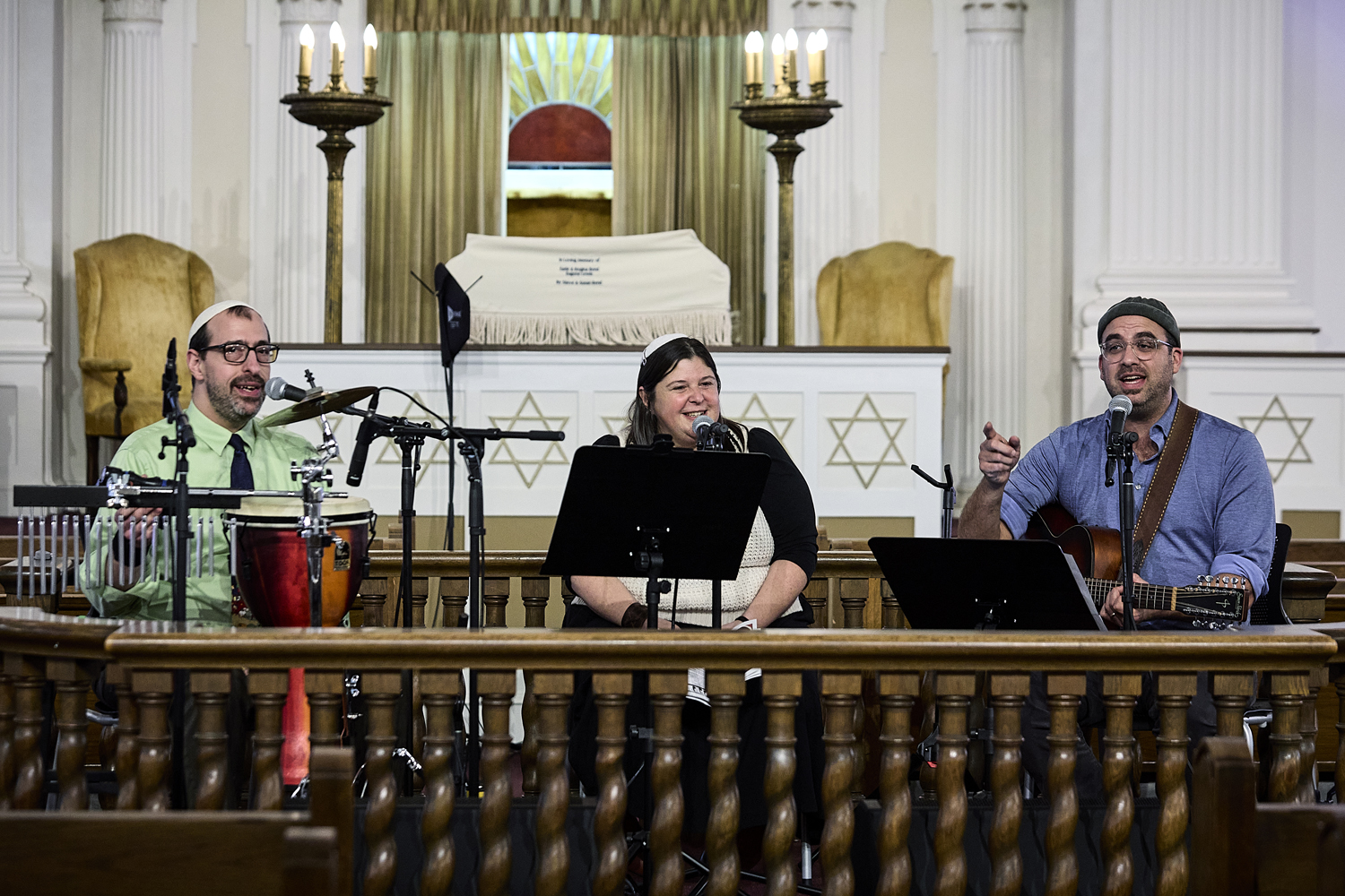 Rabbi Jessy Dressin sits between two male musicians on the stage in a synagogue, playing music.