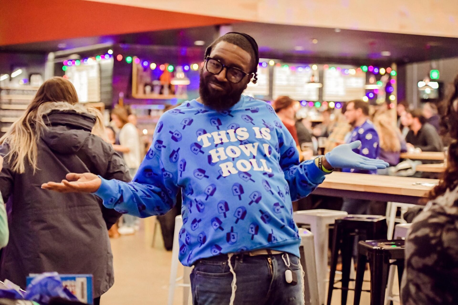 Black man wearing a blue hannukah sweaetshirt and tefillin smiles for the camera with his arms outstretched.