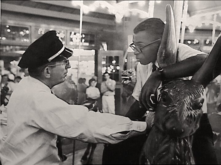 Historic, black and white photo of a white man in white shirt and dark uniform cap confronting a young black man in glasses