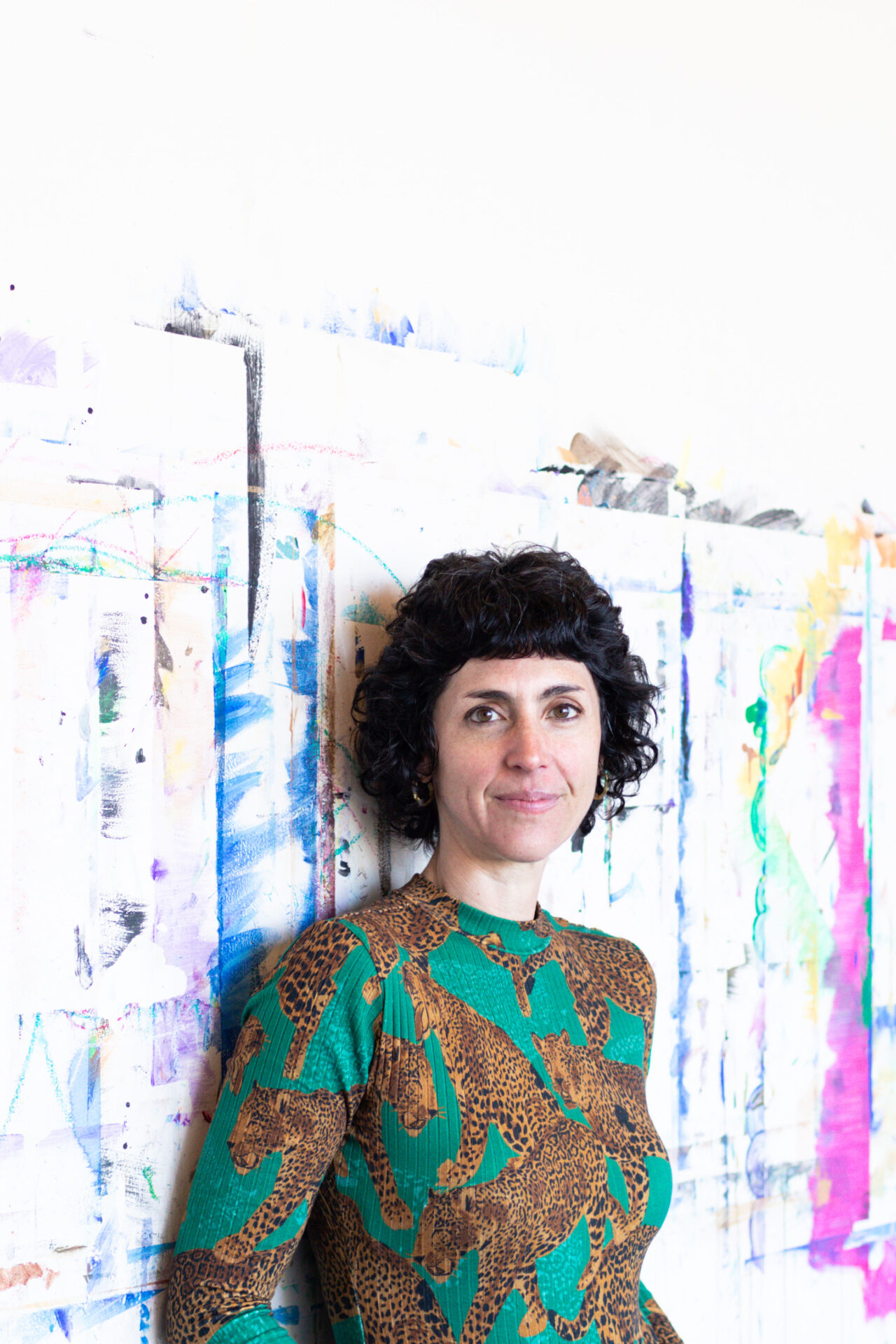 Rabbi Adina Allen, wearing a green and gold sweater, standing against a studio wall with paint remnants visible.