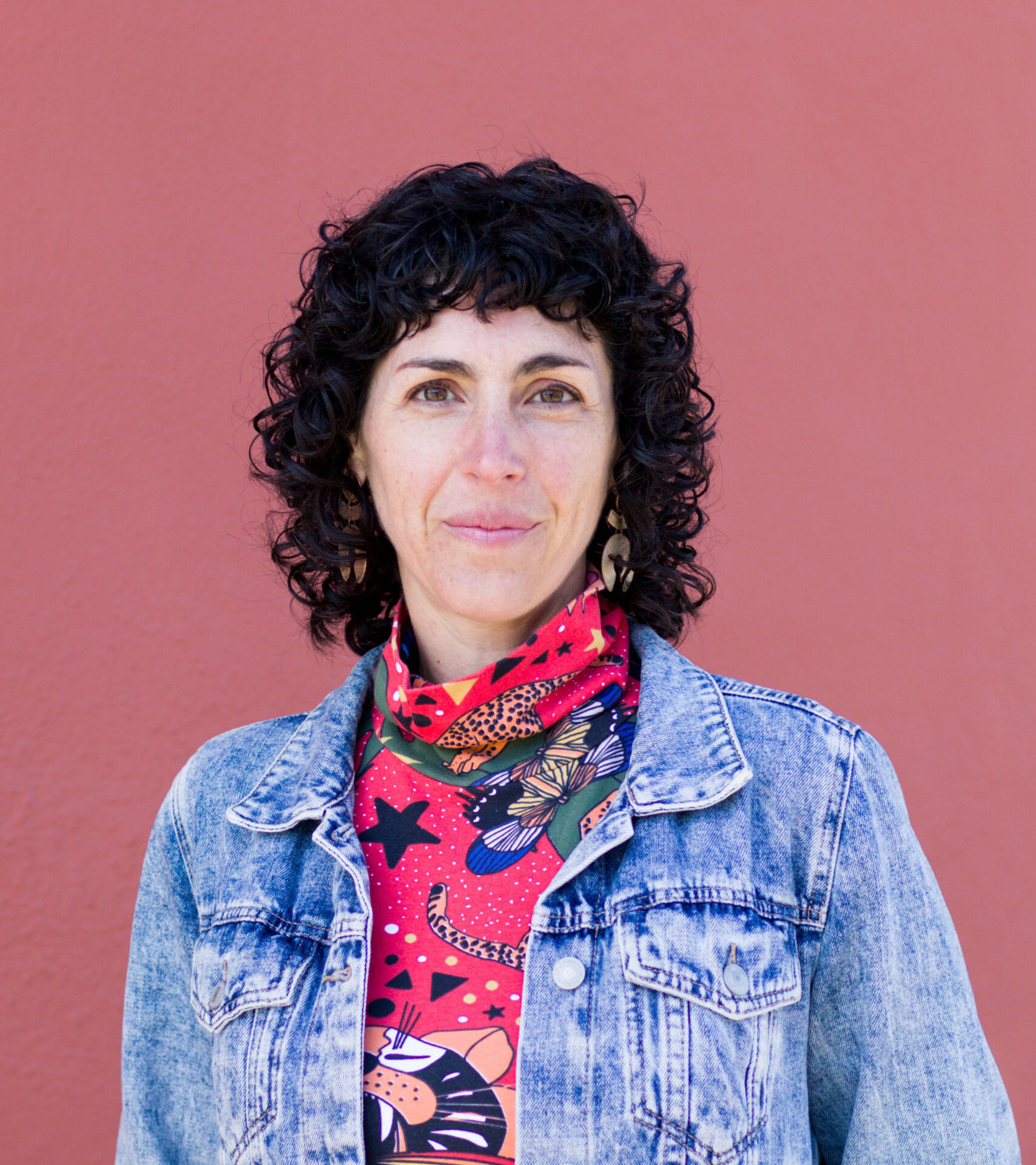 Rabbi Adina Allen, looking at the camera with curly dark hair framing her face, a pink printed scarf, and a denim jacket.