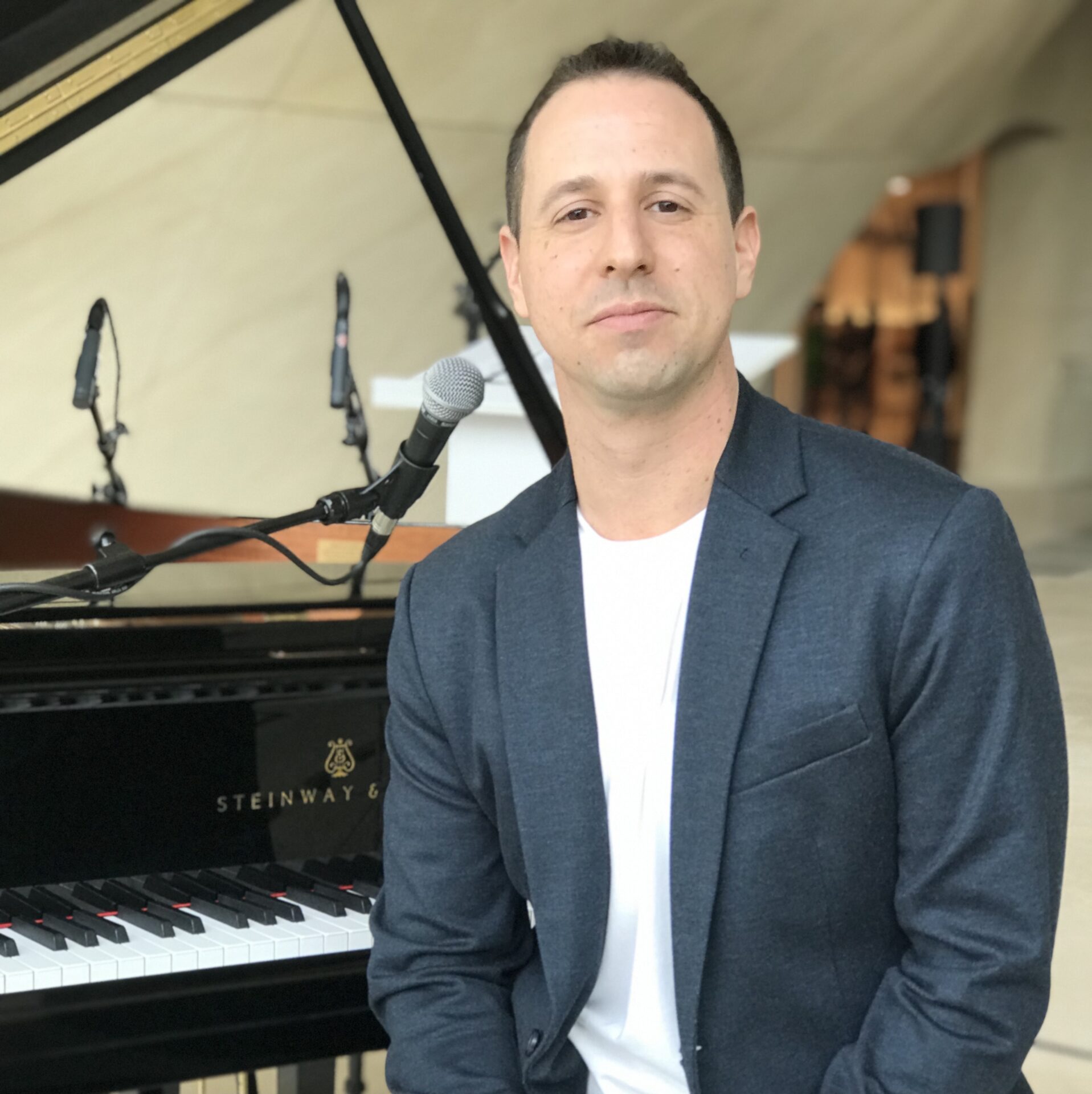 Avi Wisnia sits in front of a piano and microphone. He is wearing a tee shirt and blazer and looks directly at the camera.