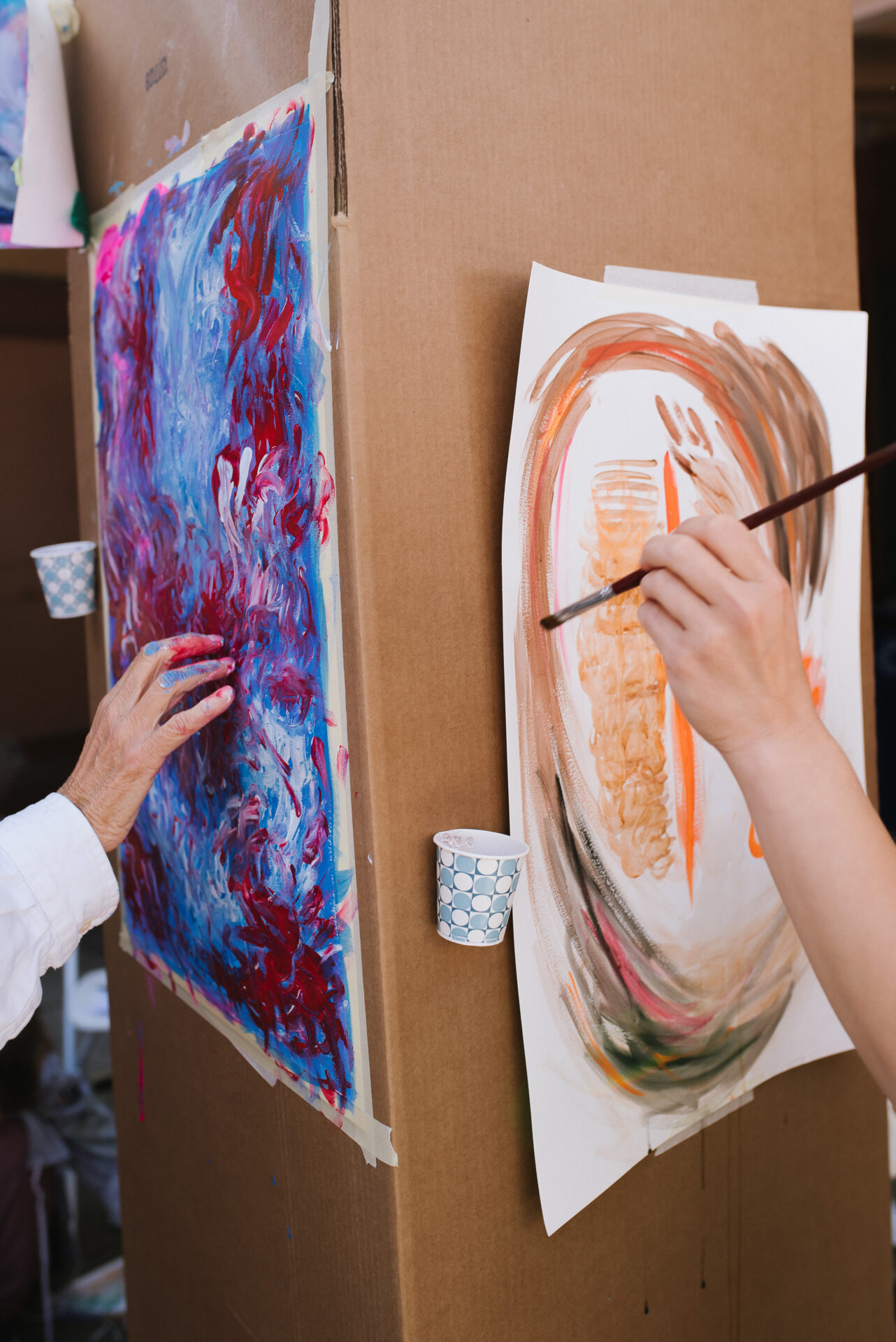 Close up of two people working on artwork tacked up on the wall. Abstract reds and blues being applied to the paper with the artist's hand on the left and brown and orange large circles being painted on the right