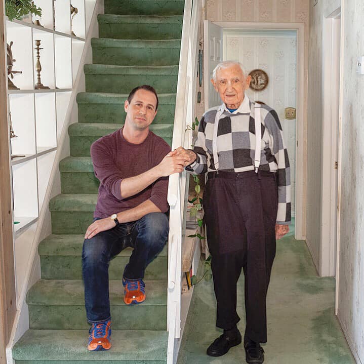 Avi Wisnia sits on a staircase on the left. His grandfather stands next to the stairs on the right. Their hands rest together on the stair railing.