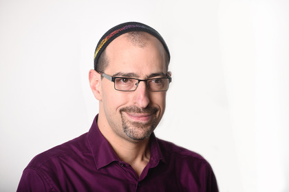 Rabbi Ilan Glazer from the shoulders up, with pale skin and close-cut hair and goatee, wearing glasses and a wide kippah and a maroon collared shirt.