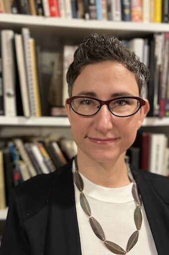 Tracie Guy-Decker, a white woman with short dark hair and glasses, looks directly at the camera. She is wearing a white blouse and dark blazer with a leaf motif necklace. There are books in the background.